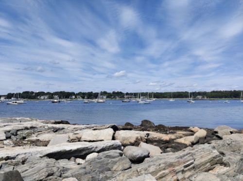 A serene coastal scene with boats anchored in the water, surrounded by rocky shores and a blue sky with clouds.