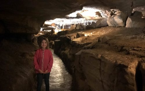 A young girl stands on a path inside a cave, with rock formations and dim lighting in the background.