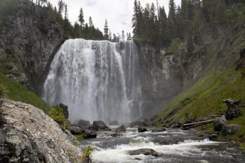 A majestic waterfall cascades down rocky cliffs, surrounded by lush greenery and tall trees under a cloudy sky.