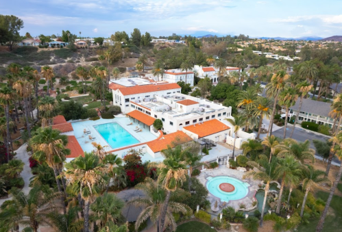 Aerial view of a resort with a large pool, palm trees, and surrounding hills in a sunny landscape.