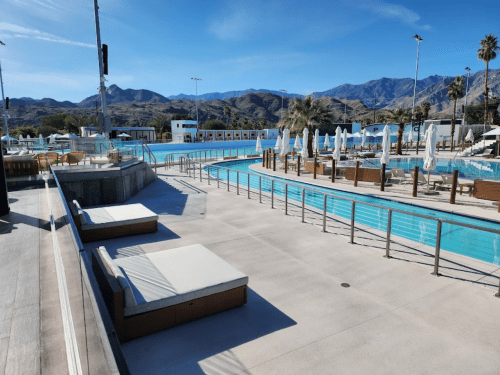 A sunny pool area with lounge chairs, surrounded by mountains and palm trees, featuring clear blue water and white umbrellas.