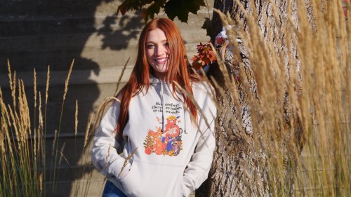 A smiling person with red hair stands by a tree, wearing a light hoodie with a colorful design, surrounded by tall grass.