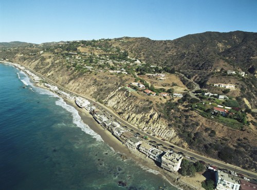 Aerial view of a coastal area with cliffs, homes, and a winding road along the beach. Clear blue sky above.