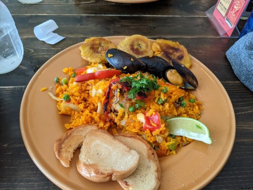 A plate of colorful rice with vegetables, mussels, fried plantains, and slices of bread, garnished with lime and herbs.