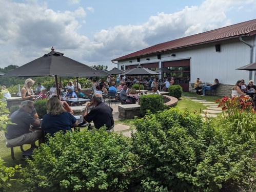 A lively outdoor dining area with people seated under umbrellas, surrounded by greenery and a rustic building in the background.