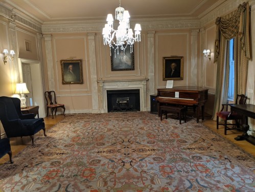 A spacious room with a chandelier, piano, ornate rug, and framed portraits on the walls. Elegant furnishings are present.