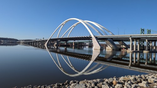 A modern bridge with arched supports spans a calm river, reflecting in the water under a clear blue sky.