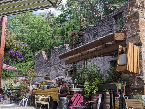 A rustic outdoor café with colorful chairs, surrounded by stone ruins and lush greenery. A vintage yellow truck is parked nearby.