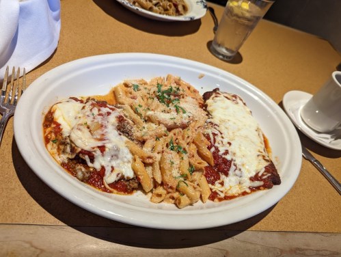 A plate of pasta with marinara sauce, cheese, and herbs, served alongside baked chicken and a glass of water.