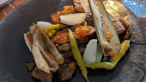 A plate of grilled seafood with vegetables, including onions, peppers, and crispy potatoes, served on a dark dish.