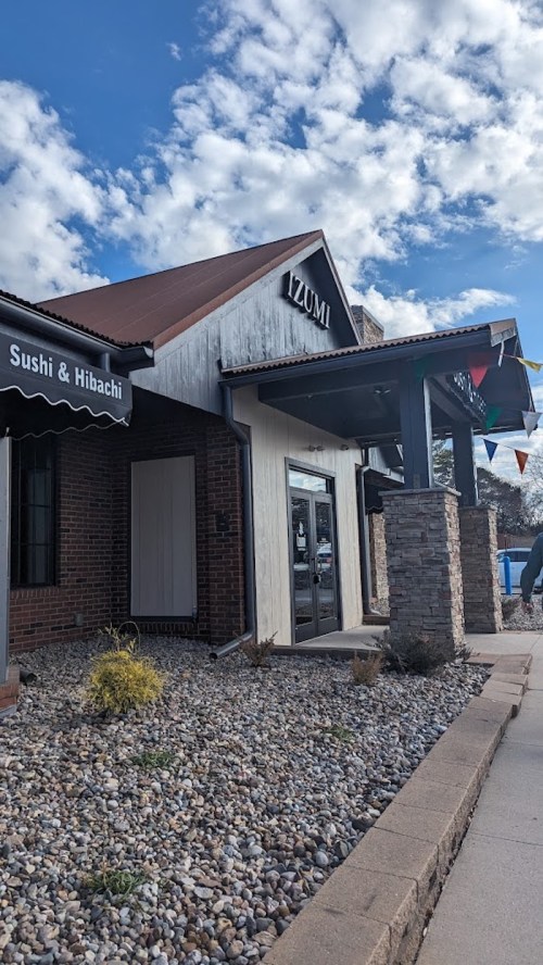 Exterior of Izumi restaurant with a stone facade, blue sky, and decorative flags. Signs for sushi and hibachi visible.