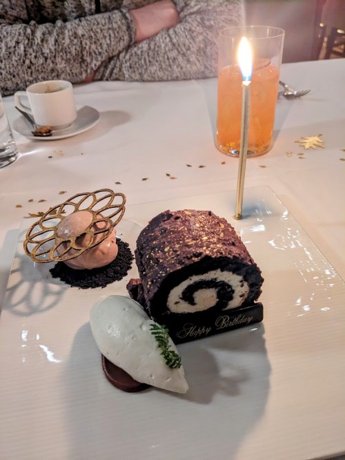 A birthday dessert featuring a chocolate roll cake, a scoop of ice cream, and a decorative candle on a white plate.