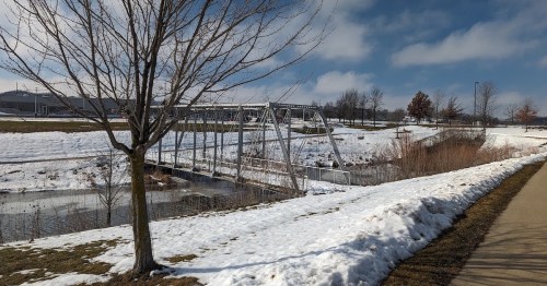 A snowy landscape featuring a bridge over a calm waterway, with bare trees and a clear blue sky in the background.