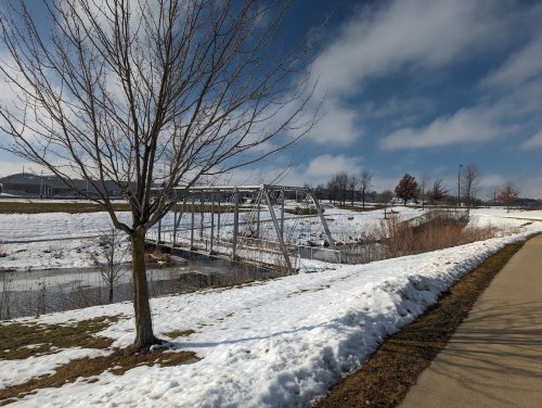 A snowy landscape with a bridge over a stream, bare trees, and a path under a blue sky with scattered clouds.