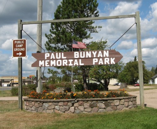 Sign for Paul Bunyan Memorial Park with an American flag, surrounded by flowers and a public campground sign.