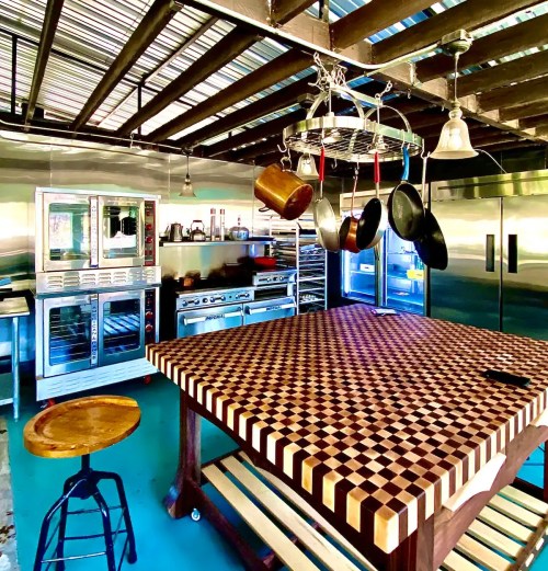 A spacious kitchen with a large checkered wooden table, stainless steel appliances, and hanging pots and pans.