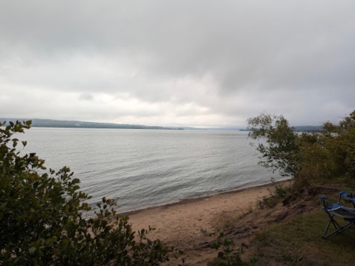A serene lakeside view with a sandy shore, surrounded by greenery under a cloudy sky.
