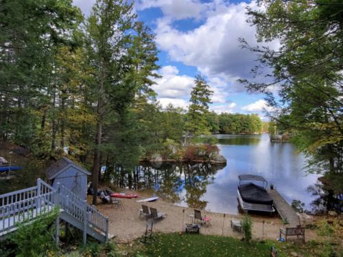 A serene lakeside view with trees, a dock, and boats under a partly cloudy sky.
