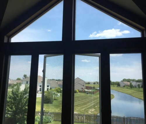 View from a large window showing a serene landscape with a pond, houses, and blue sky with clouds.