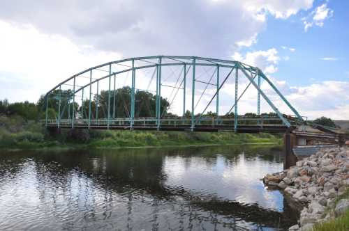 A green arch bridge spans a calm river, surrounded by lush greenery and a cloudy sky.