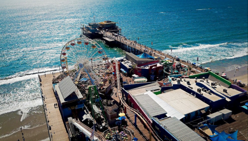 Aerial view of a vibrant pier with a ferris wheel, colorful buildings, and waves lapping at the shore.