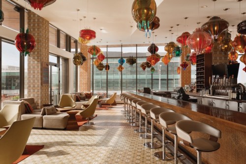 A stylish bar area with colorful hanging lights, modern seating, and large windows offering natural light.