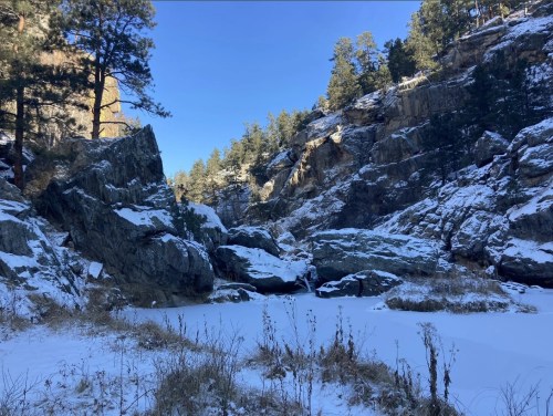 Snow-covered rocks and a frozen pond surrounded by pine trees and rocky cliffs under a clear blue sky.