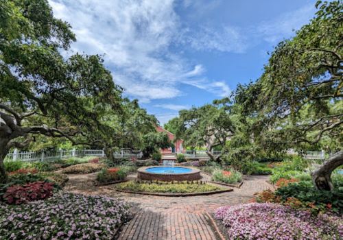 A serene garden with colorful flowers, a circular fountain, and lush trees under a blue sky.