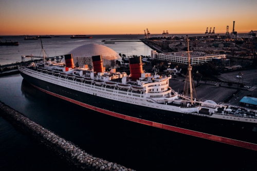 Aerial view of a historic ship docked at sunset, with a modern dome structure and cranes in the background.