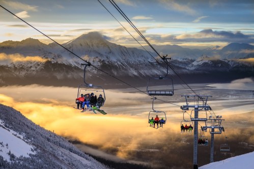 Skiers on a chairlift ascend a mountain, surrounded by clouds and stunning sunset views over a valley.