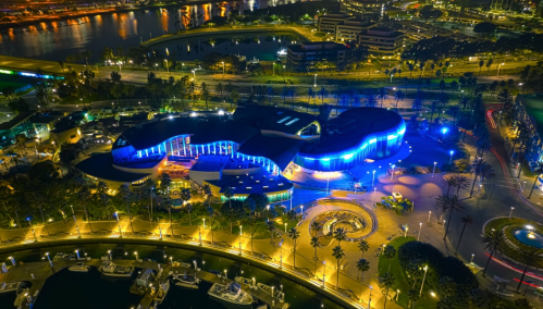 Aerial view of a modern building illuminated in blue lights, surrounded by palm trees and water at night.