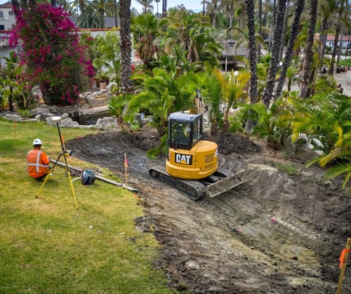 A construction site with a CAT excavator and a worker measuring land among palm trees and vibrant flowers.