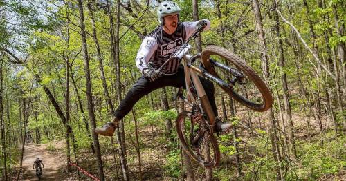 A mountain biker performs a jump over a trail in a wooded area, showcasing skill and excitement.