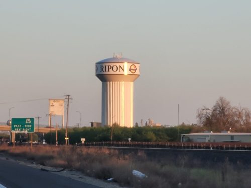 A water tower with "RIPON" written on it, surrounded by trees and a clear sky, seen from a roadside.