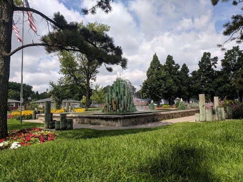 A landscaped park featuring a fountain surrounded by colorful flowers and trees under a partly cloudy sky.