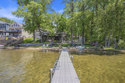 A wooden dock extends into a clear lake, surrounded by trees and houses, with people enjoying the waterfront.