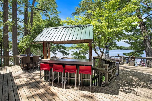 A lakeside bar with red stools, surrounded by trees and a wooden deck, under a blue sky.