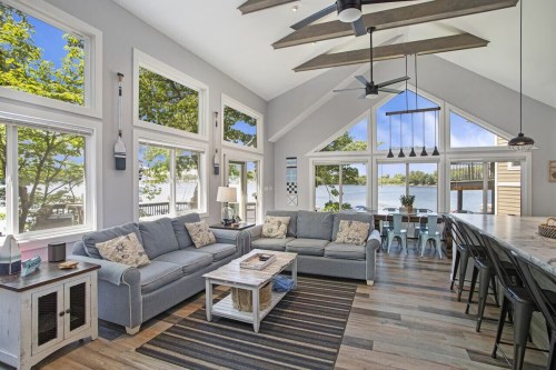Bright, modern living room with large windows, lake view, gray sofas, and a dining area featuring a long table.