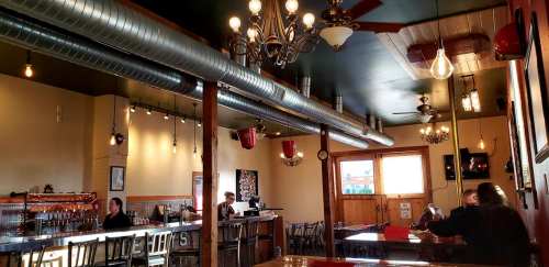 Interior of a restaurant with wooden tables, a bar, and industrial-style lighting. Patrons are seated and a server is visible.