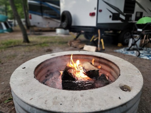 A campfire burns in a metal fire pit, with a camper and outdoor seating visible in the background.