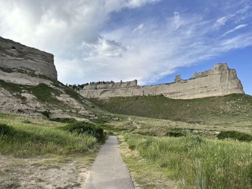 A winding path leads through lush green grass towards towering rock formations under a partly cloudy sky.