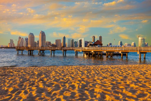 A sandy beach with a pier extending into the water, overlooking a city skyline under a colorful sunset.
