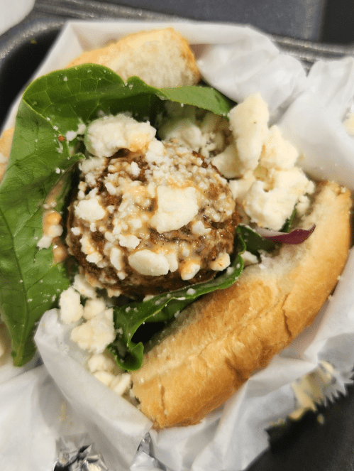 A sandwich with a meat patty, fresh greens, and crumbled cheese, served in a soft bun.