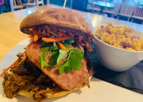 A hearty sandwich with pulled meat, topped with carrots and cilantro, served with a side of corn salad.