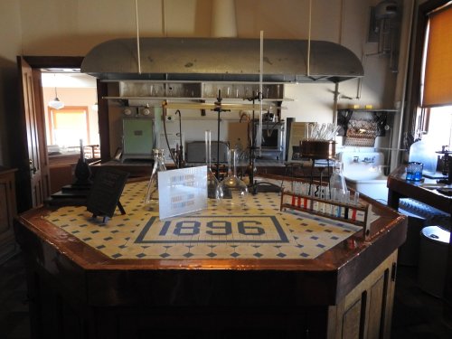 A vintage laboratory with glassware and equipment on a tiled table marked "1896." Natural light from windows.
