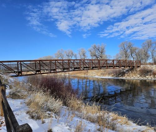 A metal bridge spans a calm river, surrounded by trees and snow under a blue sky with scattered clouds.