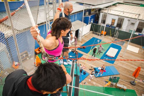 A group of climbers prepares to descend from a high indoor climbing structure, with mats and equipment below.