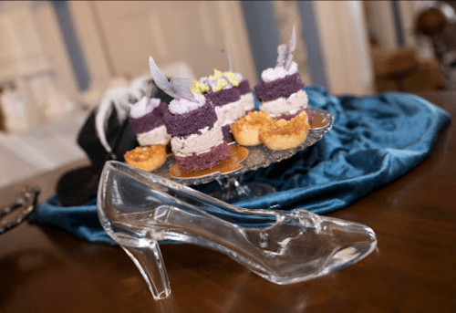 A clear glass high heel shoe beside a tiered dessert display with purple and white cakes and small pastries.