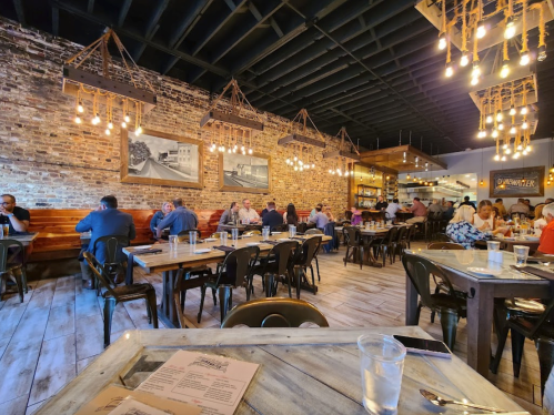 A bustling restaurant interior with wooden tables, exposed brick walls, and hanging lights, filled with diners.