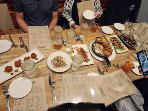 A table filled with various dishes, drinks, and menus, with people seated around enjoying a meal together.